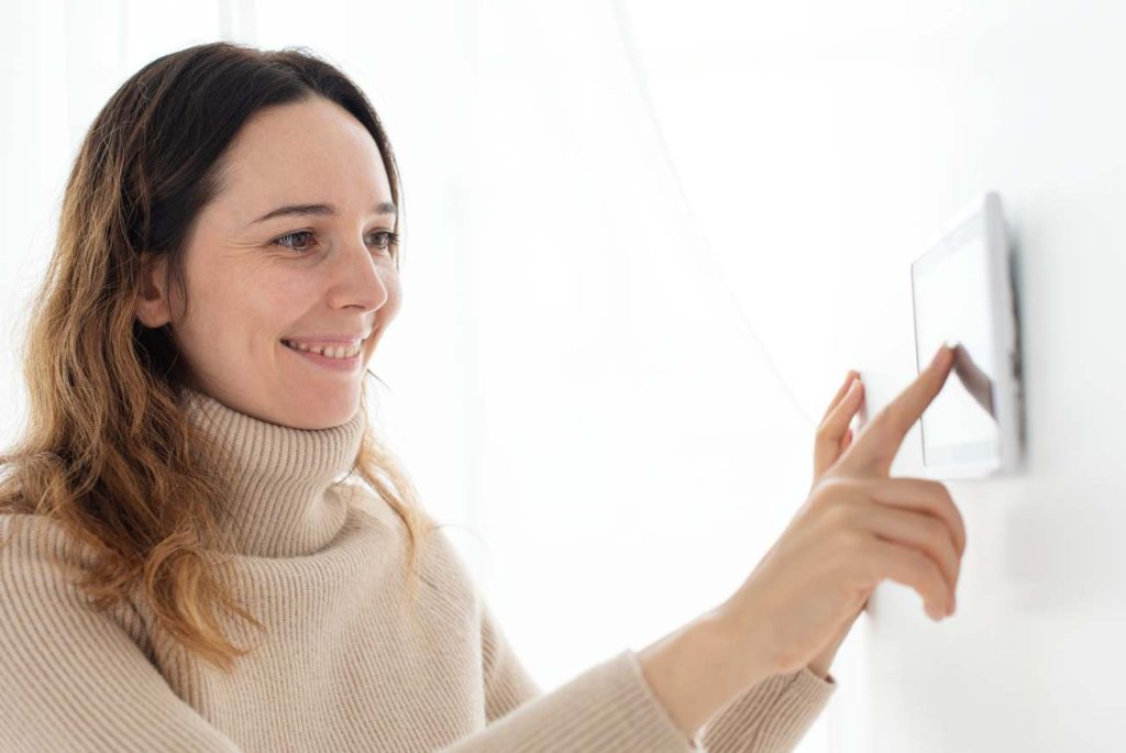 Une femme souriante vêtue d'un pull beige ajuste le réglage idéal thermostat chauffage hiver sur un boîtier mural blanc.